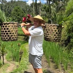Derek carrying two woven baskets on a bamboo pole through a rice paddy, in a conical hat